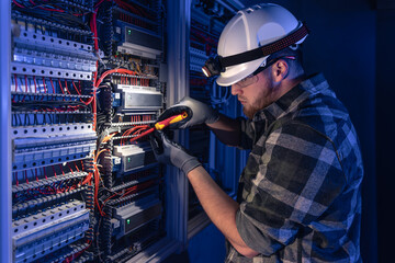 A male electrician works in a switchboard in overalls against the backdrop of emergency lighting.