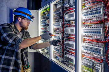A male electrician works in a switchboard in overalls against the backdrop of emergency lighting.