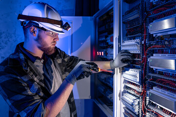 A male electrician works in a switchboard in overalls against the backdrop of emergency lighting.