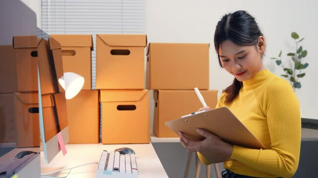 Asian woman checking inventory and writing on clipboard, surrounded by cardboard boxes in her office