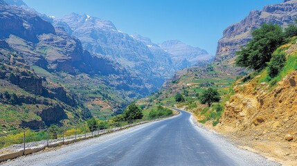 Naklejka premium Winding road through a picturesque mountain valley, surrounded by lush greenery and rugged terrain under a clear sky.