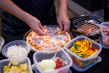 Chef hands is putting sliced pineapples on Hawaiian pizza with ingredients in boxes on a black table.