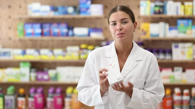 Kind young apothecary holding a box with remedy standing against shelves full of medicine in drugstore