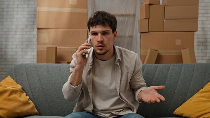Young couple moving into new apartment, space filled with cardboard boxes, man sits on the sofa in room of new apartment and talks on smartphone, frustrated negative expression.