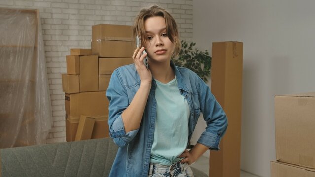 Young couple moving into new apartment, space filled with cardboard boxes, woman stands in the room of new flat and talks on smartphone, frustrated negative expression.