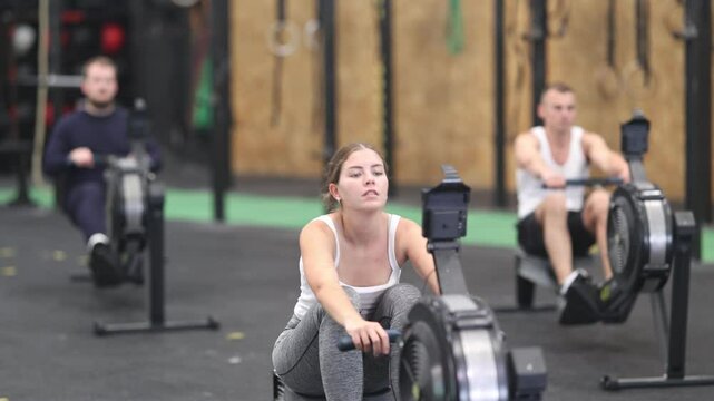 Emotional sporty young girl doing functional strength training, working out on rowing machine in modern gym
