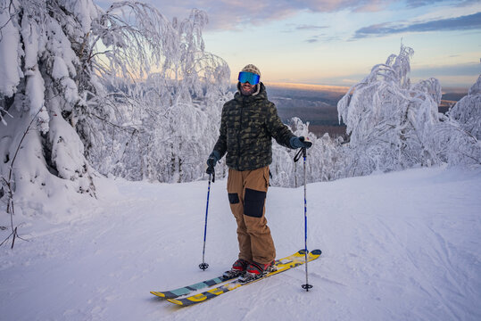 Skier stands on a snowy slope with ski poles, enjoying the view of a beautiful winter landscape at sunrise, surrounded by snow-covered trees