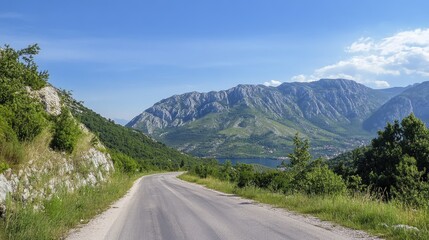 Naklejka premium Scenic asphalt road winding through lush green mountains with clear blue sky and distant hills in picturesque landscape view