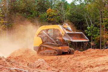 Skid steer loader works on clearing earth amid trees, creating dusty environment during preparing...