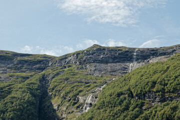 Waterfalls gracefully flow over rugged cliffs, surrounded by lush greenery under a bright sky.