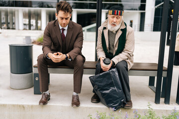 Front view of focused business man in formal attire attentively engages in conversation with senior homeless man holding disposable coffee cup, while seated on bench in public area, on cloudy day.