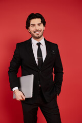 Confident young man in business attire holding a laptop against a vibrant red backdrop