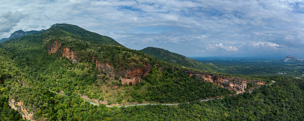 Aerial panoramic view of lush green hills and valleys intersected by a winding mountain road. The landscape features rugged cliffs, dense forests, and a distant horizon under a partly cloudy sky.