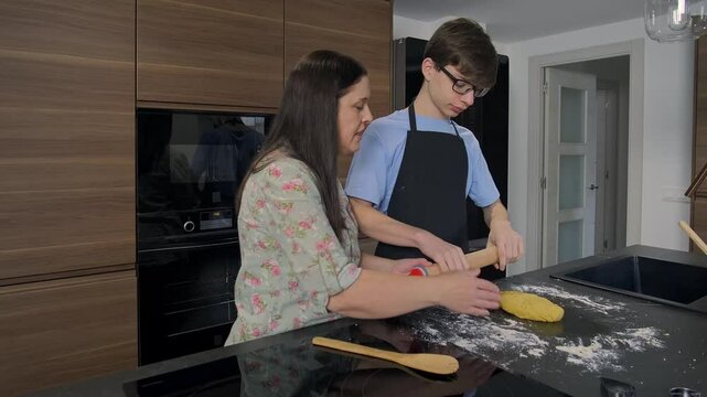 Mother and teenage son bond in the kitchen, learning to bake together. Ingredients and tools surround them to create meal, sharing love and communication. Woman helping child with rolling pin cooking.