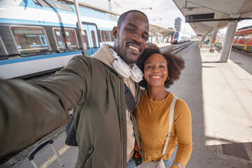 Joyful African American couple taking a selfie at a railway station before embarking on their train journey together. Travel happiness concept.