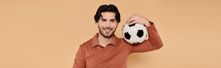 Handsome young man with soccer ball poses cheerfully against warm background