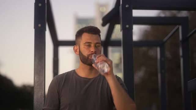 Athletic man drinking water from a bottle after outdoor training session. Refreshing hydration during a workout in the urban park at dusk.