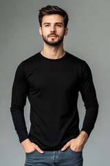 Obraz premium A young man in a black long sleeve T-shirt mockup stands confidently with hands in pockets against a gray background. He has a well-groomed beard and a serious expression.