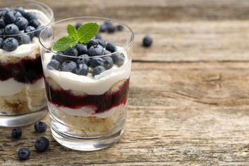 Tasty trifle dessert. Sponge cake, blueberries, jam and whipped cream in glasses on wooden table, closeup. Space for text