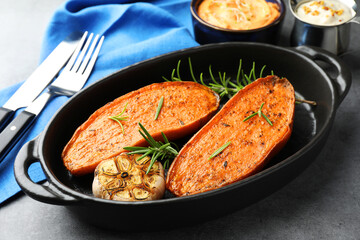 Halves of tasty cooked sweet potato with rosemary and garlic in baking dish served on grey table, closeup
