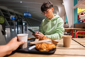 Woman using smartphone and eating croissant at airport cafe