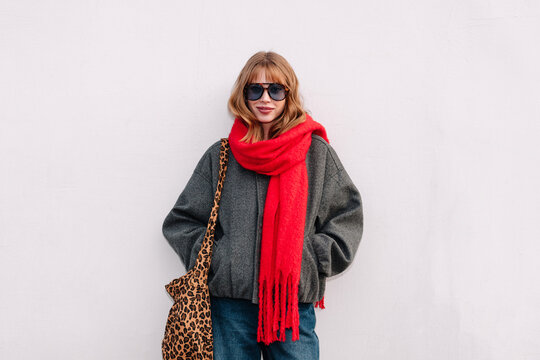 Woman in sunglasses and a red scarf stands against a wall