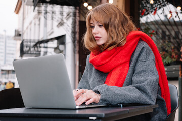 A woman in a red scarf works on her laptop at an outdoor cafe