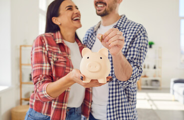 Happy young family couple with financially stable life saving up money together. Cheerful smiling married man and woman put coins inside a piggy bank and have fun together. Finance, budget concept