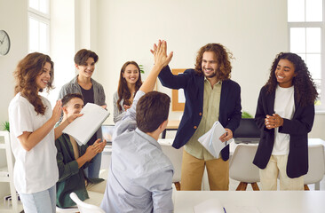 Young friendly male teacher and happy high school students giving high five to each other in the classroom rejoicing the success in studying. Back to school, education, support and teamwork concept.