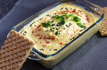 Homemade chickpea hummus, sprinkled with parsley, red paprika and black sesame seeds, is on the table in a glass transparent form, next to two dry diet bread.