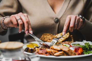 A close-up of a person enjoying a delicious meal, featuring assorted meats, vegetables, and bread on plate. hands hold a knife and fork, ready to savor the feast. A cozy dining atmosphere is captured.