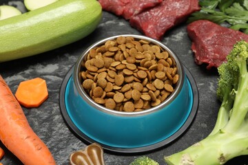 Dry pet food in feeding bowl and natural products on dark textured table, closeup