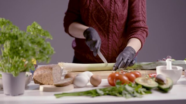 Krojenie pora na r&oacute;wne kawałki ostrym nożem na drewnianej desce. Obok widoczne są świeże warzywa, takie jak pomidory, awokado, seler naciowy, jajka i pieczywo, tworząc apetyczne tło