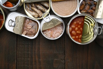 Open tin cans with different preserved products on wooden table, flat lay. Space for text