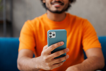 Young man wear orange casual clothes sits on blue sofa couch hold use mobile cell phone stay at...