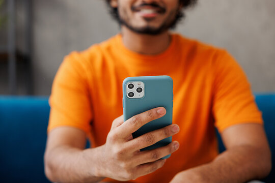 Young man wear orange casual clothes sits on blue sofa couch hold use mobile cell phone stay at home hotel flat rest relax spend free spare time in living room indoor. Lounge concept. Focus on device.