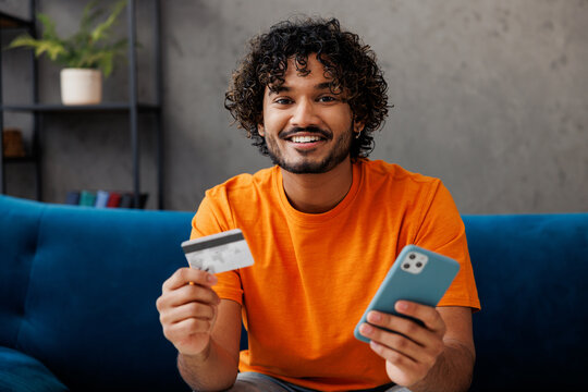 Young man wear casual clothes sit on blue sofa couch use mobile cell phone credit bank card shopping online stay home hotel flat rest relax spend free spare time in living room indoor. Lounge concept