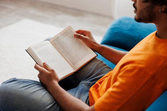 Close up side view young Indian man wears orange casual clothes sits on blue sofa couch read novel book stay at home hotel flat rest relax spend free spare time in living room indoor. Lounge concept.