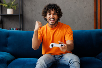 Young Indian man wear orange casual clothes sit on blue sofa couch hold play pc game with joystick...