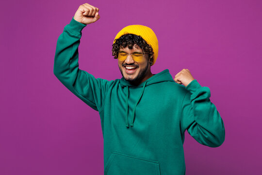 Young overjoyed excited happy cool Indian man he wear green hoody yellow hat sunglasses casual clothes do winner gesture dance isolated on plain purple background studio portrait. Lifestyle concept.