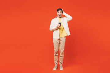 Full body young surprised amazed impressed winner man he wear white shirt yellow t-shirt casual clothes hold in hand use mobile cell phone isolated on plain red orange background. Lifestyle concept.
