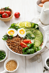 Buckwheat groats with avocado, various vegetables and soft-boiled egg in a ceramic bowl on a white table, close up view. Healthy and delicious vegetarian food
