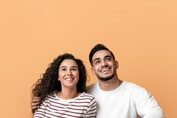 Fototapeta premium Dreamy Young Arab Couple Sitting On Floor And Looking Up At Copy Space, Pensive Smiling Middle Eastern Spouses Thinking About Interesting Offer While Posing Over Yellow Studio Background