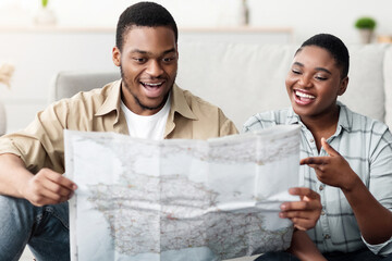 Family Vacation. Excited African American Couple Holding Touristic Map Choosing Travel Destination Sitting Near Unpacked Suitcase At Home. Tourism And Traveling Concept Selective Focus