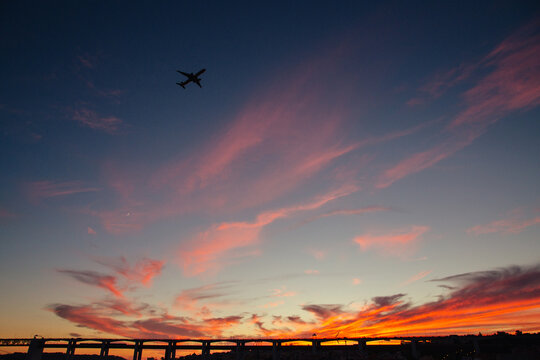 Airplane flying over a vibrant sunset sky near a city bridge