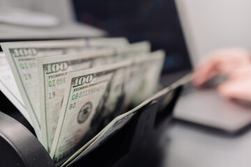 Fototapeta premium Close-up of counting 100 dollar bills on a financial professional's desk next to a laptop computer in a counting machine. Concept of banks, loans, deposits or currency exchange and stocks.