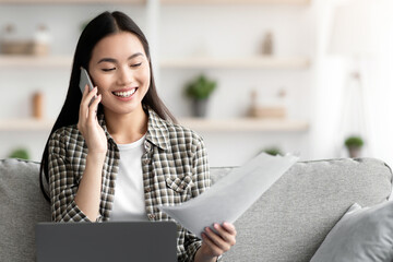 Pretty young asian woman having phone conversation, sitting on couch in living room and using laptop, looking at documents. Cheerful lady working from home, websurfing and talking on mobile phone