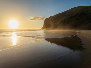 Obraz premium Golden hour sunset over a tranquil beach. Coastal cliffs reflect in the calm water. Peaceful scenery. Anawhata, Piha, Auckland, New Zealand