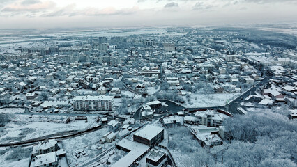 Aerial View of Snow-Covered Town in Winter. Wide aerial view of a serene town blanketed in snow, featuring rooftops, roads, and surrounding fields under a cloudy winter sky.