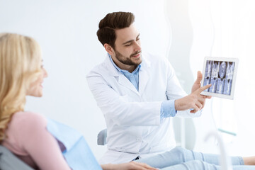 Obraz premium Male dentist showing female patient treatment goal, using digital tablet, dental clinic interior. Bearded man stomatologist having conversation with lady in dental chair, pointing at gadget